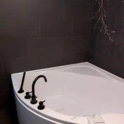 Biała narożna wanna z czarną armaturą na tle ciemnych płytek i dekoracyjnej ściany z motywem gałęzi; na wannie leżą zapakowane elementy sterujące.