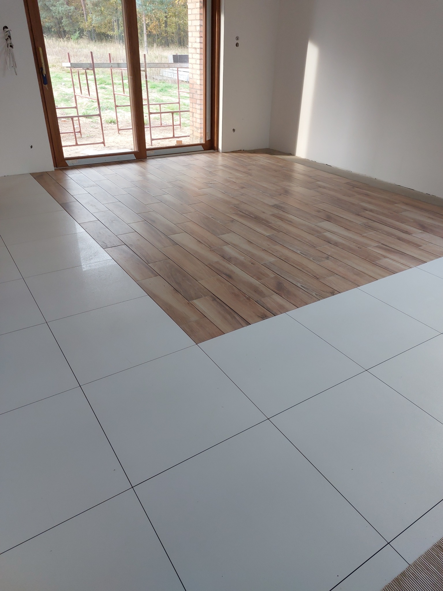 Układanie podłogi: połączenie białych, kwadratowych płytek i paneli imitujących drewno w jasnym wnętrzu z dużym oknem balkonowym.