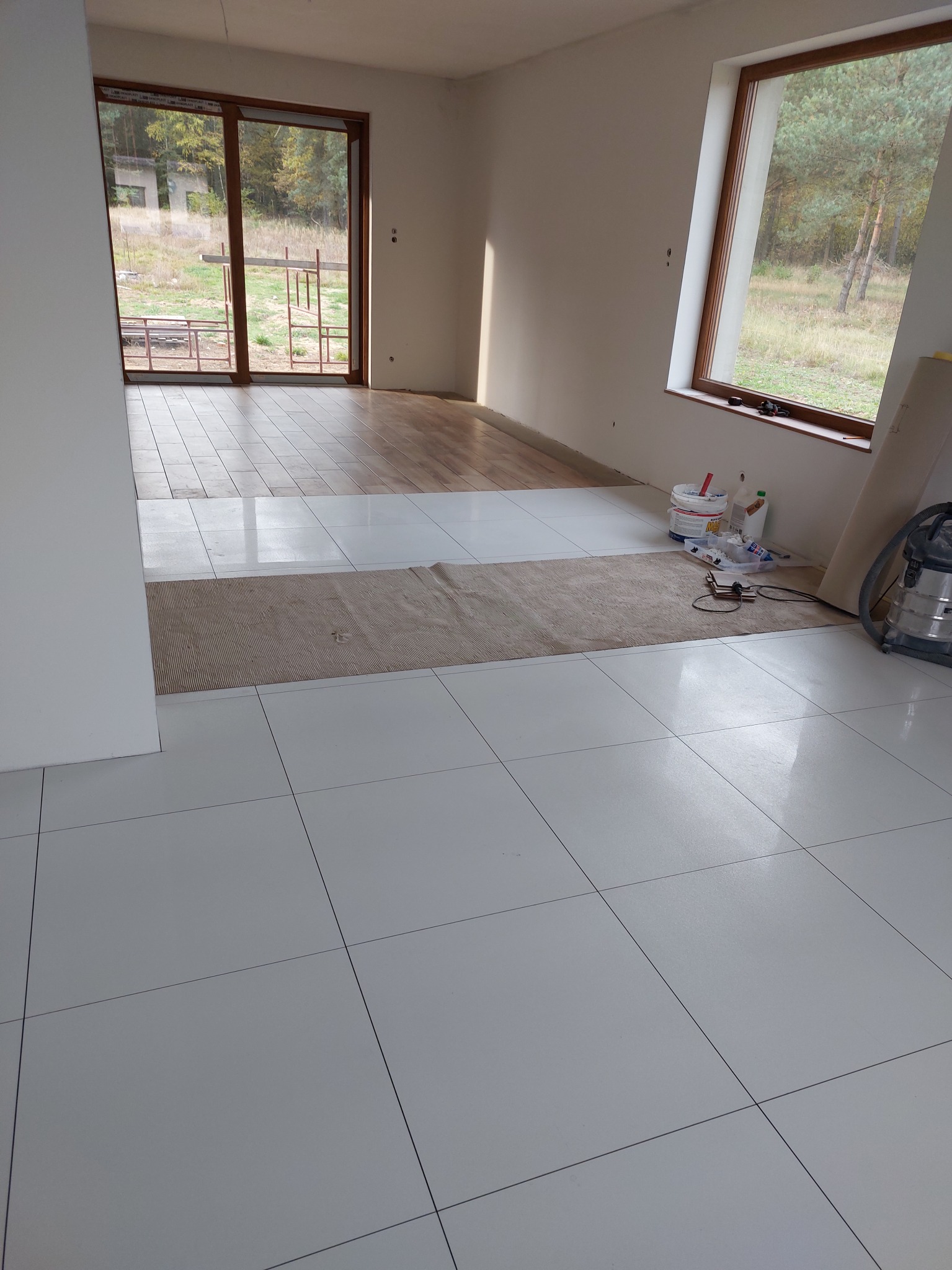 Wnętrze w trakcie prac wykończeniowych: białe płytki podłogowe, fragment parkietu, okno z widokiem na zewnątrz, narzędzia i materiały budowlane przy ścianie.