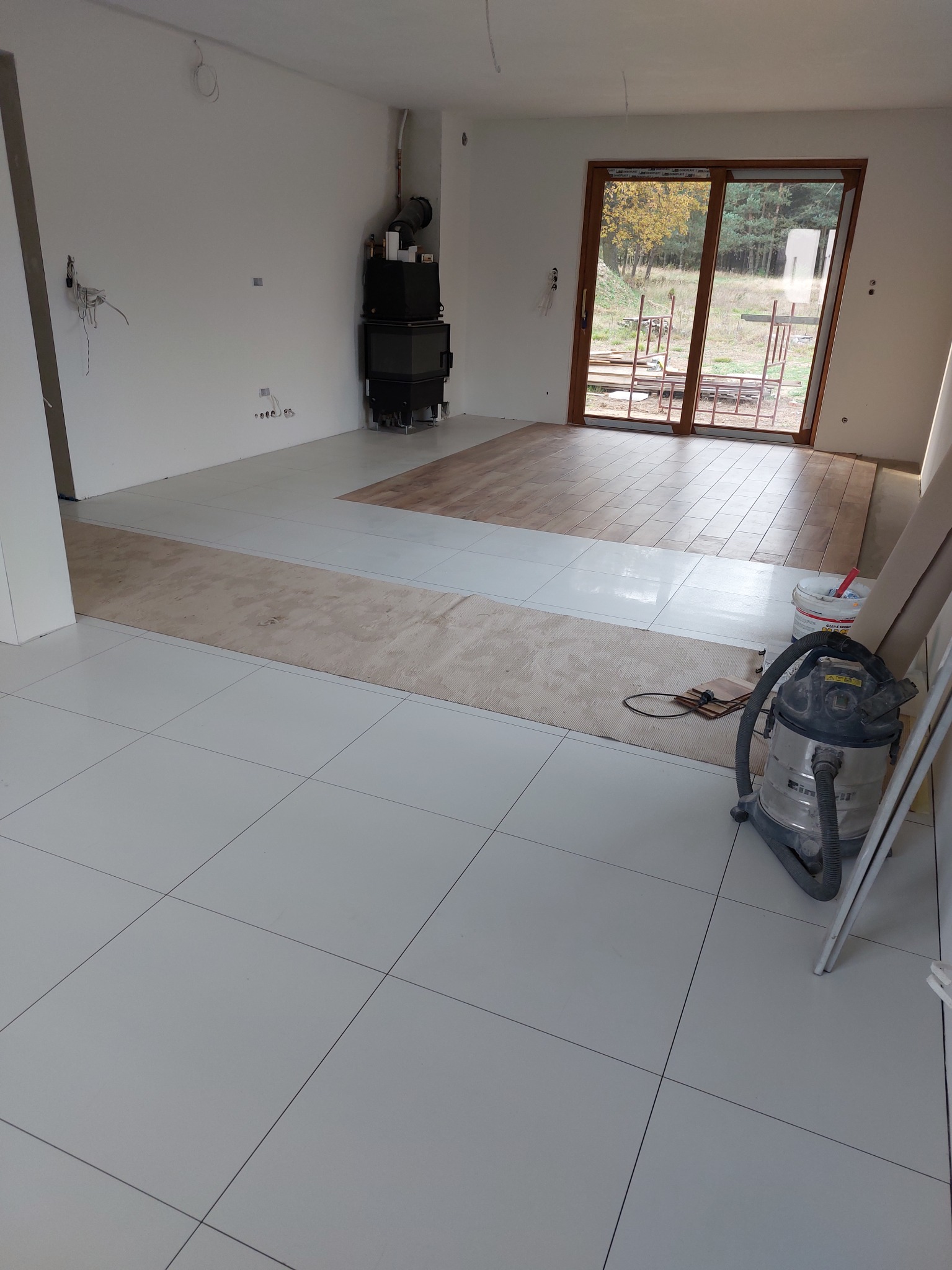 Wnętrze w trakcie prac wykończeniowych: świeżo ułożone białe płytki podłogowe, fragment podłogi z paneli, kominek, okno balkonowe z widokiem na zewnątrz, odkurzacz przemysłowy i materiały budowlane.