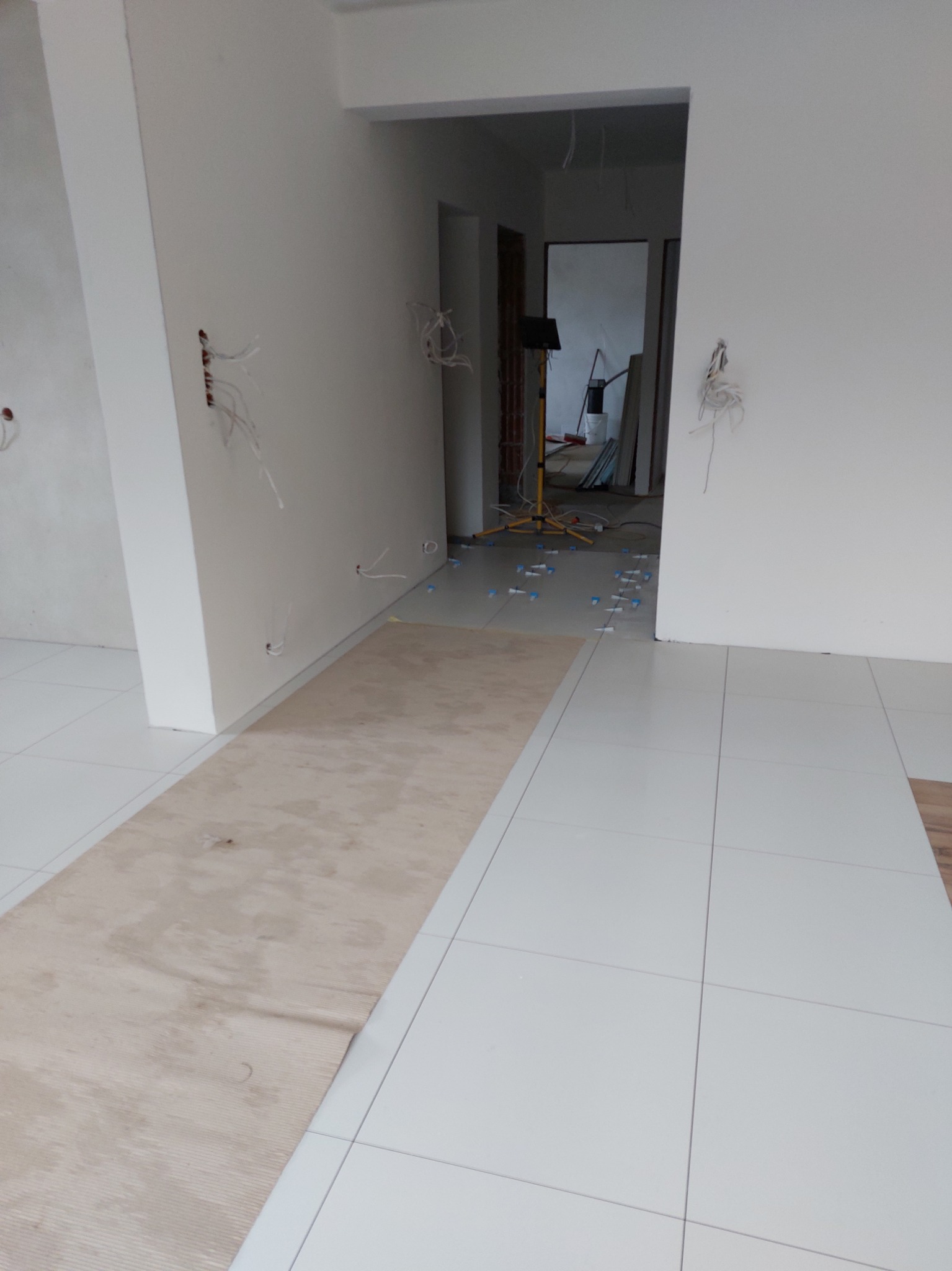 Białe płytki podłogowe w trakcie układania, z widocznymi krzyżykami dystansowymi i zabezpieczającą tekturą, ściany przygotowane do malowania z wystającymi kablami elektrycznymi.