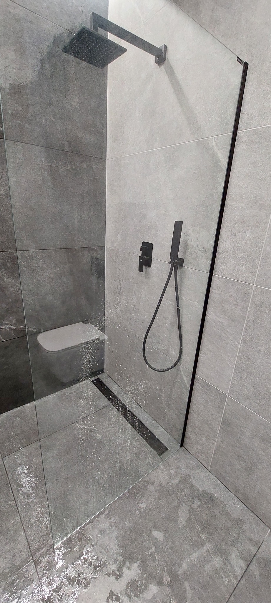 Nowoczesna kabina prysznicowa walk-in z czarną armaturą i odpływem liniowym, wykończona szarymi płytkami imitującymi kamień.