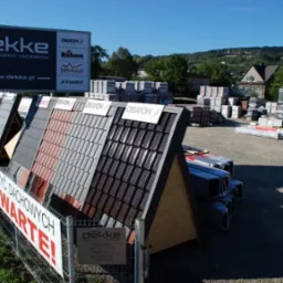 Ekspozycja różnych wzorów i kolorów pokryć dachowych na stojakach przed sklepem Dekke, widoczne palety z materiałami budowlanymi i tablica reklamowa z logo producentów.