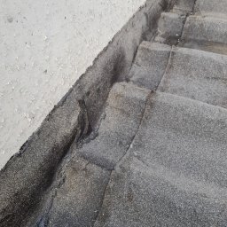 Dah - Fragment dachu pokrytego papą z widocznymi łączeniami i uszkodzeniami, przylegający do ściany z łuszczącą się farbą.