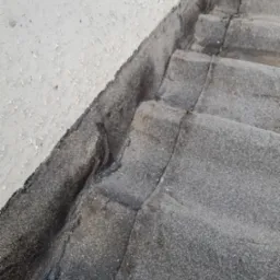 Fragment dachu pokrytego papą z widocznymi łączeniami i uszkodzeniami, przylegający do ściany z łuszczącą się farbą.