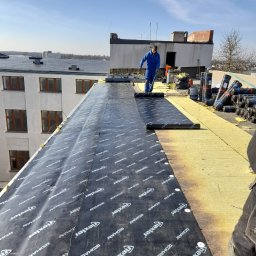 Dah - Prace na płaskim dachu budynku: dekarz w niebieskim kombinezonie rozprowadza papę termozgrzewalną z widocznym logo 'Texler' na izolacji z wełny mineralnej, w tle budynki mieszkalne i błękitne niebo.