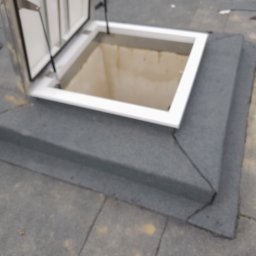 Dah - Otwarty świetlik dachowy z białą ramą na pokrytym papą dachu płaskim, widoczne detale obróbki blacharskiej wokół podstawy świetlika.