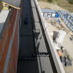 Montaż izolacji termicznej na dachu budynku, widoczna butla gazowa i palnik dekarski, w tle kontenery budowlane.