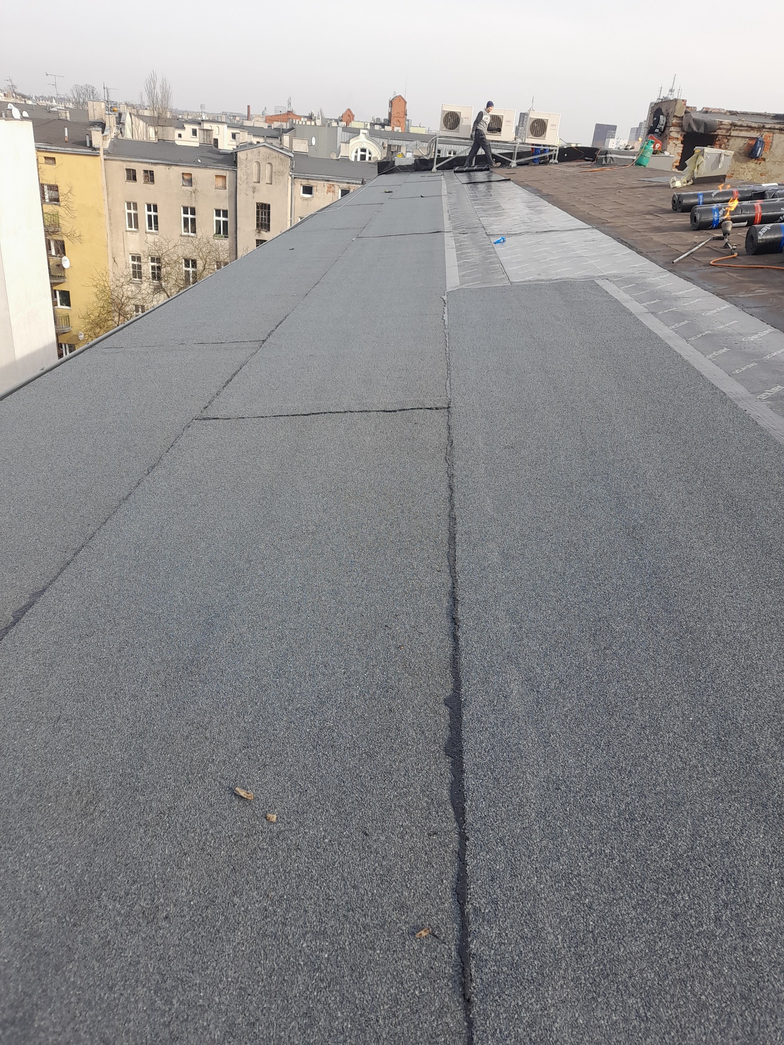 Prace na dachu płaskim pokrytym papą termozgrzewalną, widoczne zgrzewane pasy papy, pracownicy w oddali i urządzenia klimatyzacyjne na dachu. W tle zabudowa miejska.