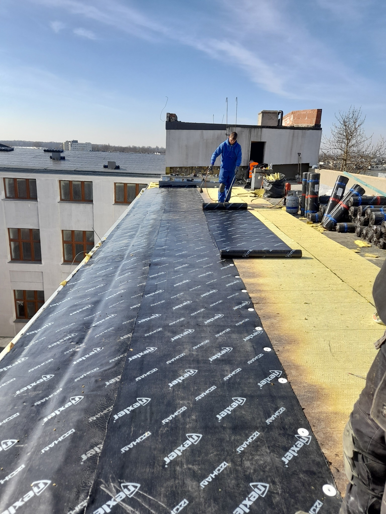 Prace na płaskim dachu budynku: dekarz w niebieskim kombinezonie rozprowadza papę termozgrzewalną z widocznym logo 'Texler' na izolacji z wełny mineralnej, w tle budynki mieszkalne i błękitne niebo.