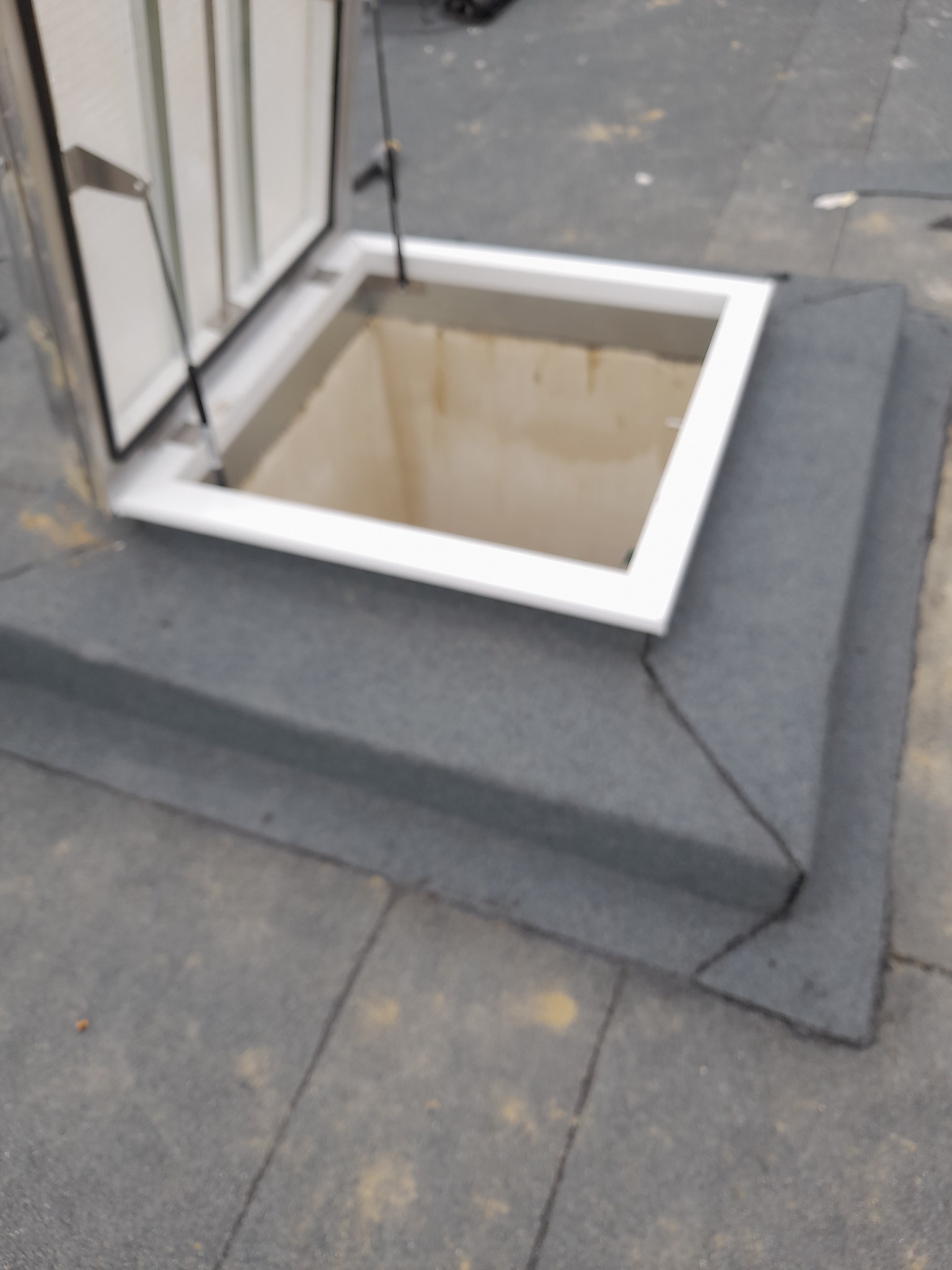 Otwarty świetlik dachowy z białą ramą na pokrytym papą dachu płaskim, widoczne detale obróbki blacharskiej wokół podstawy świetlika.