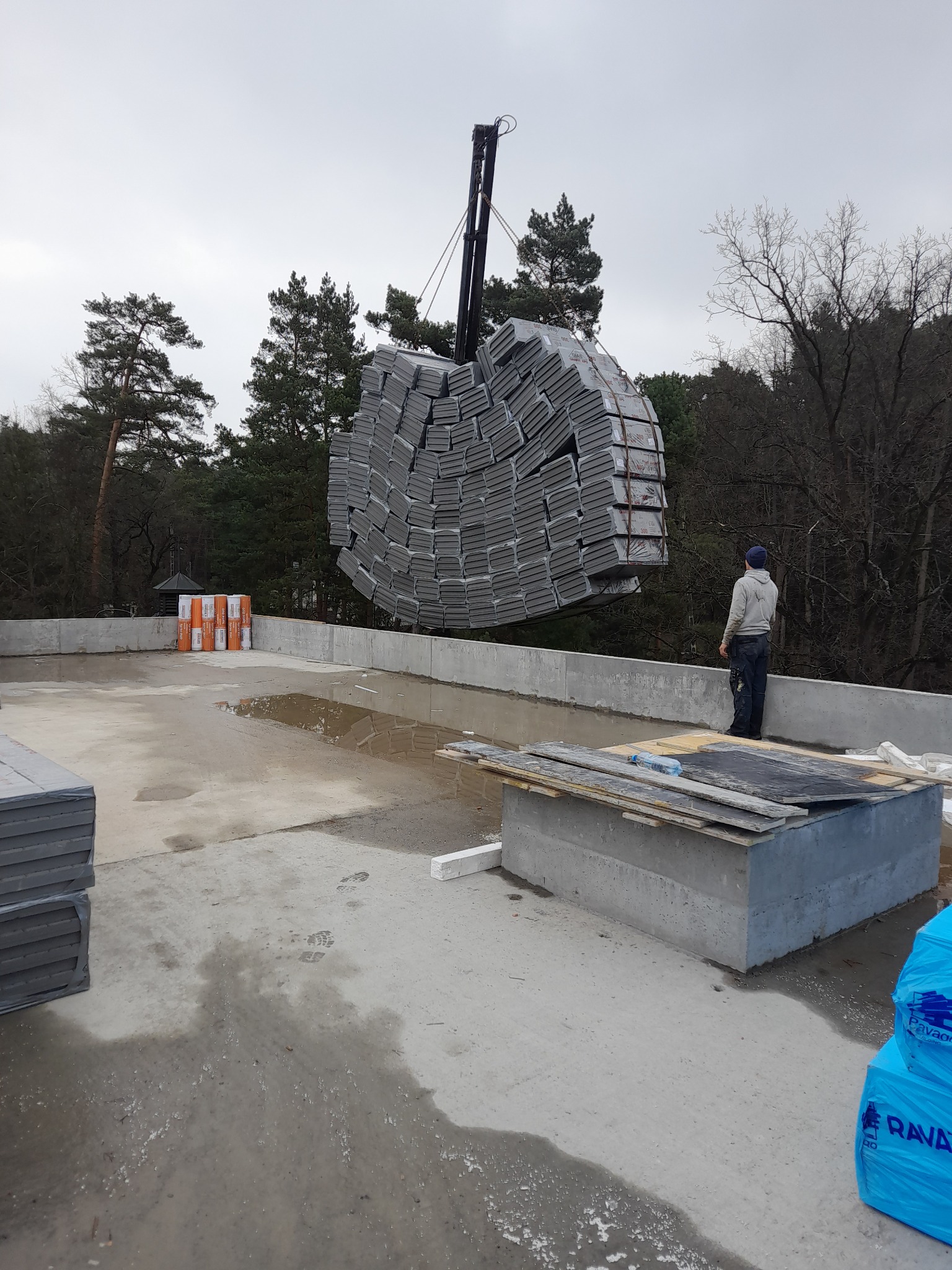 Pracownik obserwuje podnoszenie dźwigiem pakietu szarych płyt izolacyjnych na płaski dach budynku w trakcie budowy, widoczne pakiety materiałów budowlanych i kałuże na dachu.