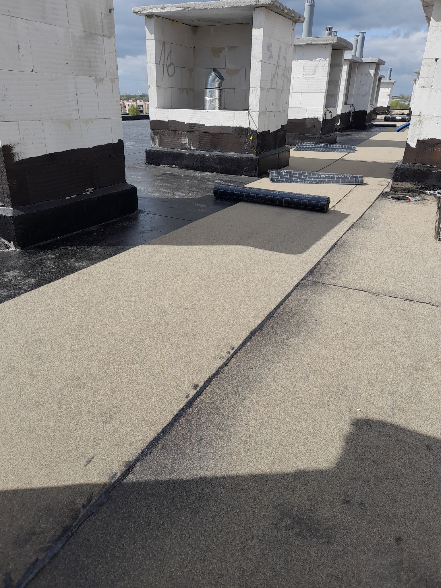 Płaski dach z widocznymi kominami wentylacyjnymi w trakcie uszczelniania, z rozłożonymi rolkami papy bitumicznej Swissopor Bikutop.