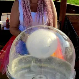 Uśmiechnięta kobieta z różowymi warkoczami stoi za maszyną do waty cukrowej marki King, na tle zielonego wzgórza i drewnianej altany.