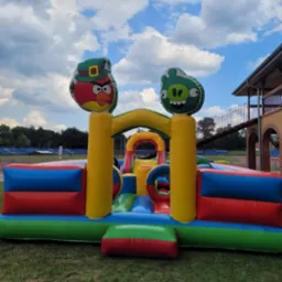 Kolorowy dmuchany zamek z motywami Angry Birds na zewnątrz, z widocznym budynkiem w tle.