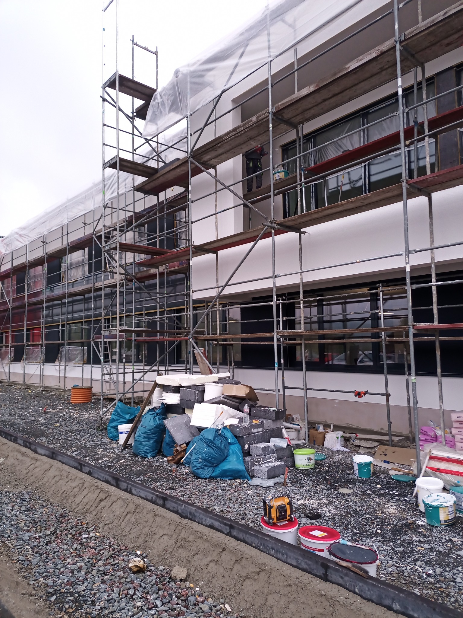 Budynek w trakcie prac elewacyjnych: rusztowania, folia ochronna, materiały budowlane i narzędzia na placu budowy, widoczny pracownik na rusztowaniu.