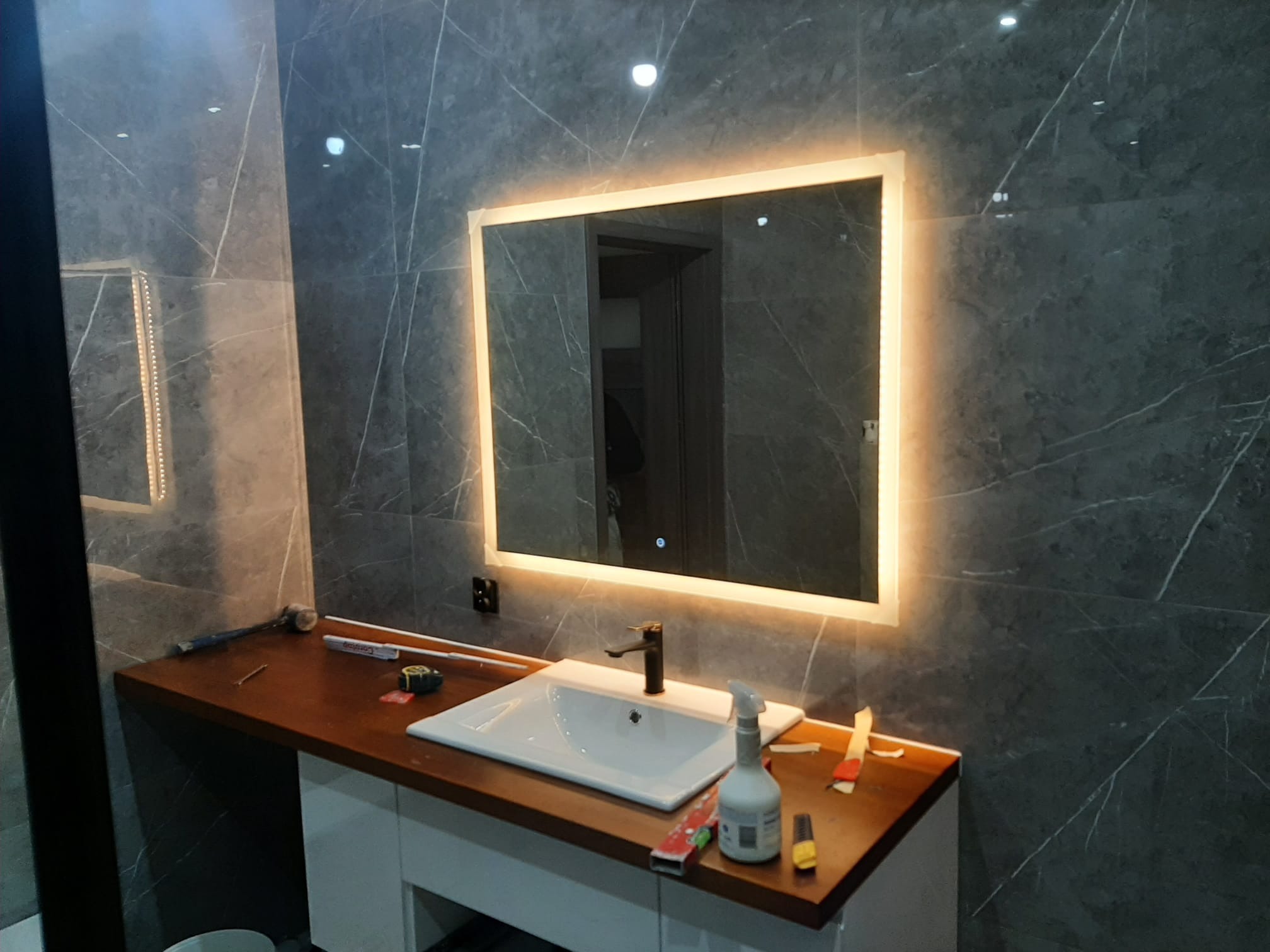 Łazienka w trakcie wykańczania: umywalka wpuszczana w drewniany blat, podświetlane lustro na szarej ścianie z marmurowym wzorem, widoczne narzędzia i akcesoria malarskie.