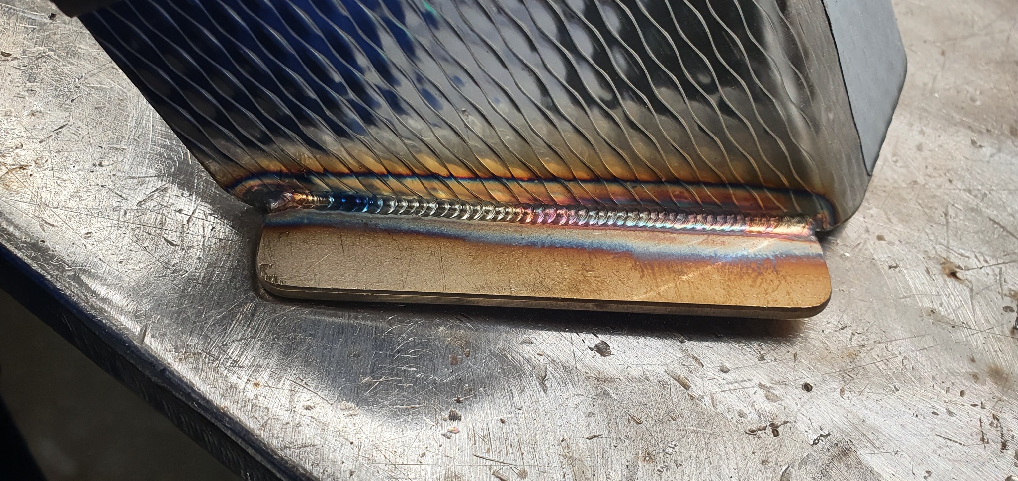 Zbliżenie na spaw łączący dwa metalowe elementy, z widocznymi kolorami hartowania i śladami obróbki na powierzchni stołu spawalniczego.