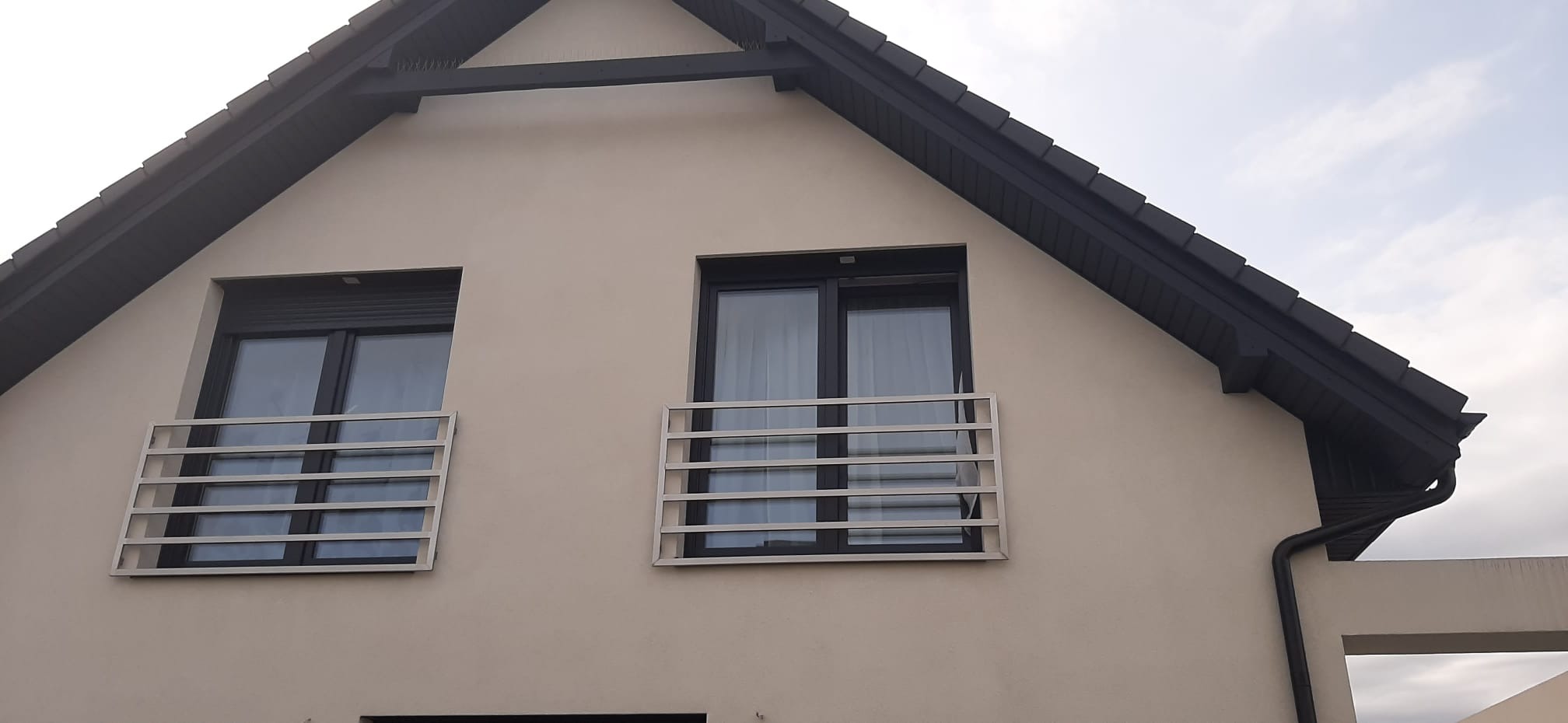Dwa okna na elewacji domu zabezpieczone zewnętrznymi, poziomymi konstrukcjami z metalowych profili.