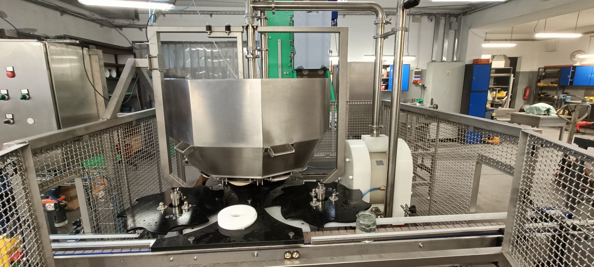 Automatyczna linia produkcyjna z metalową konstrukcją, zbiornikiem i siatkową osłoną ochronną, w tle szafki i narzędzia.