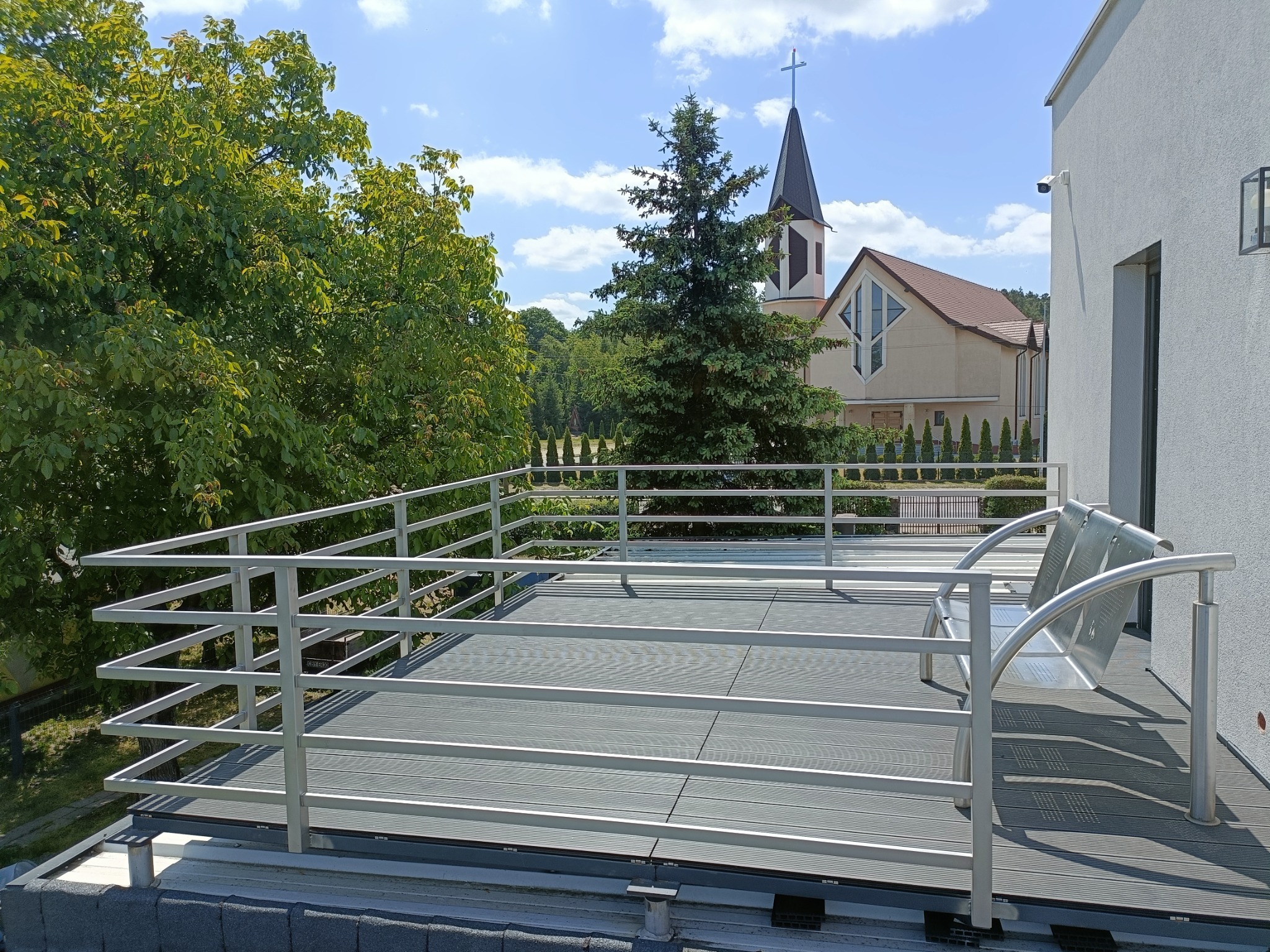 Nowoczesny taras na dachu z szarą podłogą kompozytową i metalową balustradą z widokiem na kościół i zieleń.