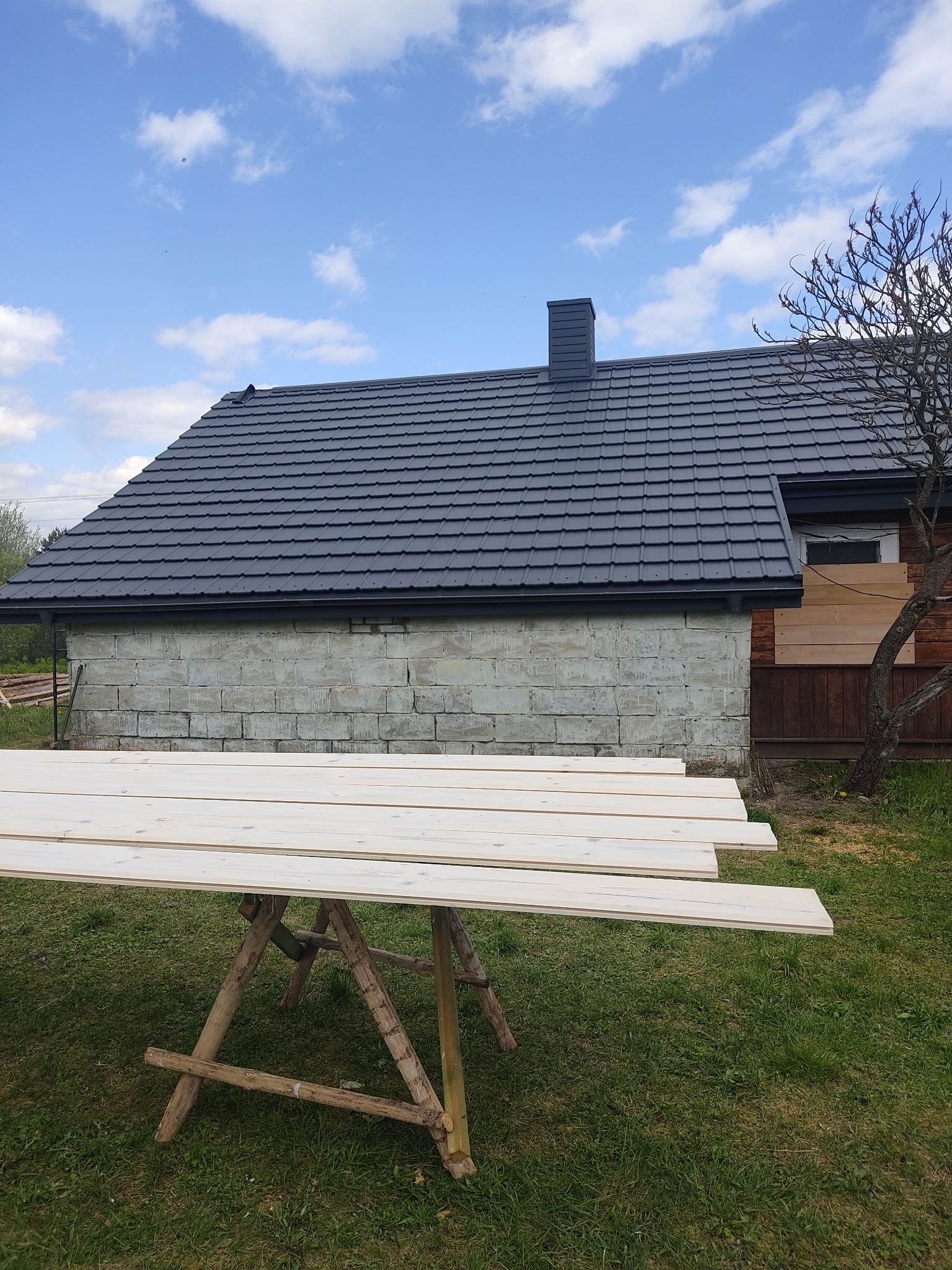 Budowa domu: ściana z bloczków, nowy dach z ciemnej dachówki, zabezpieczone okno deskami, drewniane deski na kozłach na trawie w słoneczny dzień.