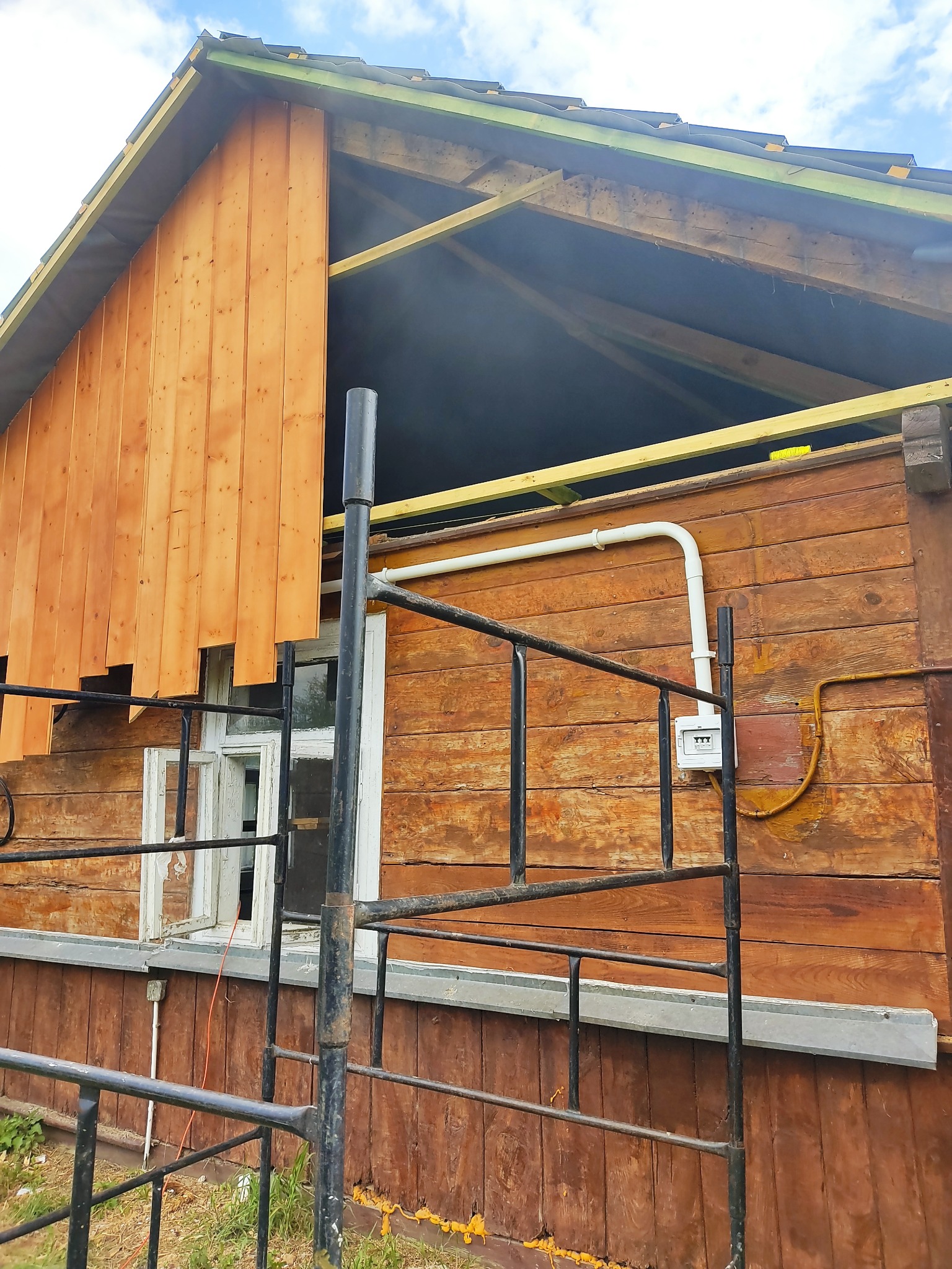 Renowacja drewnianego domu: fragment elewacji z nową, jasną boazerią kontrastujący ze starą, ciemniejszą okładziną, widoczne rusztowanie i instalacja elektryczna.