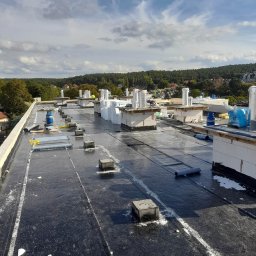 Dach Sergii Kuzmuk - Czarny, płaski dach pokryty papą termozgrzewalną w trakcie prac, widoczne kominy wentylacyjne zabezpieczone folią, butla gazowa, rolki papy, bloczki betonowe, w tle zieleń drzew i zabudowania.