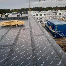 Dach Sergii Kuzmuk - Widok z góry na płaski dach pokryty czarną membraną z widocznym logo producenta, na dachu leżą palety z drewnem, w tle budynki w trakcie budowy i żurawie budowlane.