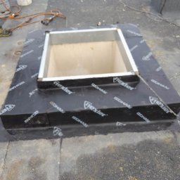 Dach Sergii Kuzmuk - Montaż obróbki blacharskiej wokół otworu w dachu płaskim, zabezpieczonego membraną z nadrukiem, widoczne narzędzia i materiały budowlane w tle.