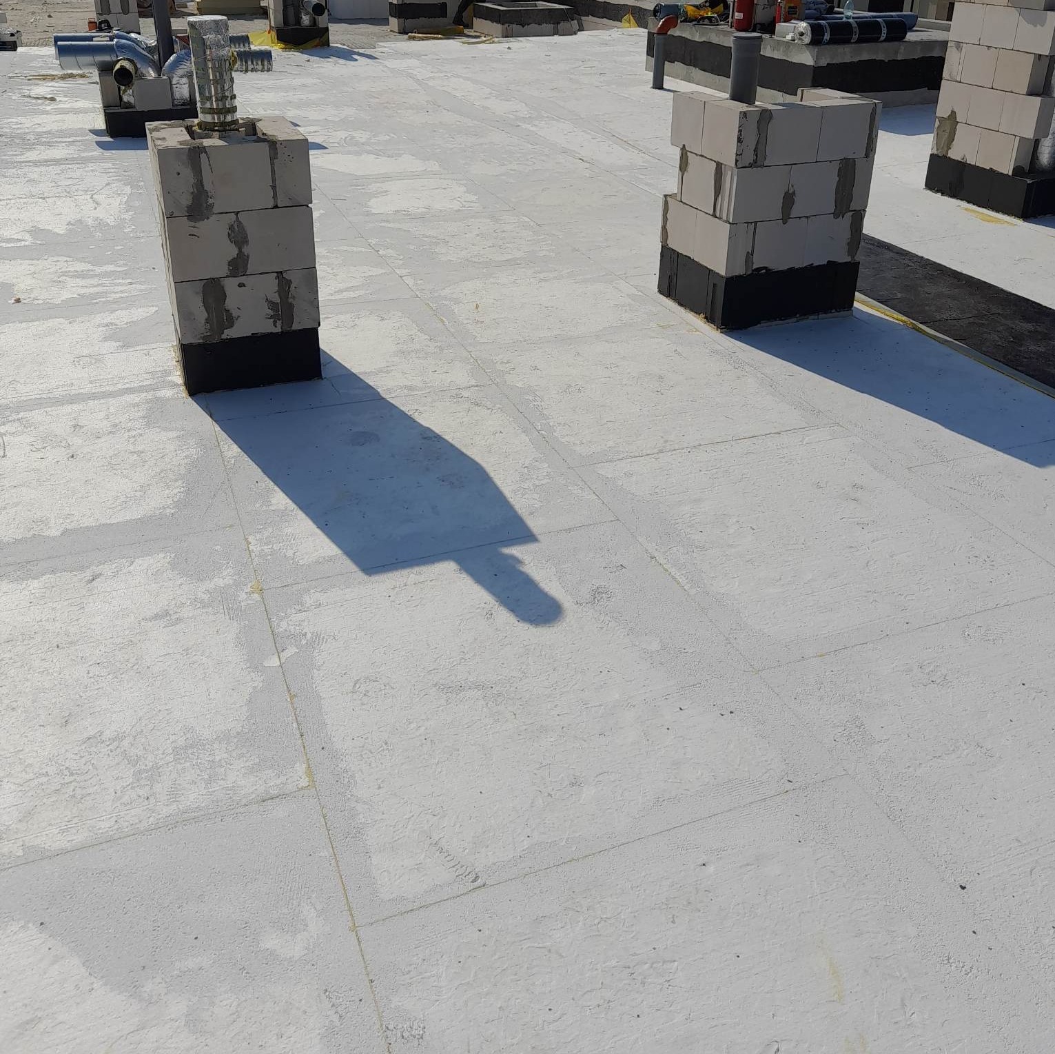 Płaski dach pokryty białą membraną z widocznymi kominami wentylacyjnymi obudowanymi bloczkami betonowymi, zabezpieczonymi czarną izolacją u podstawy.