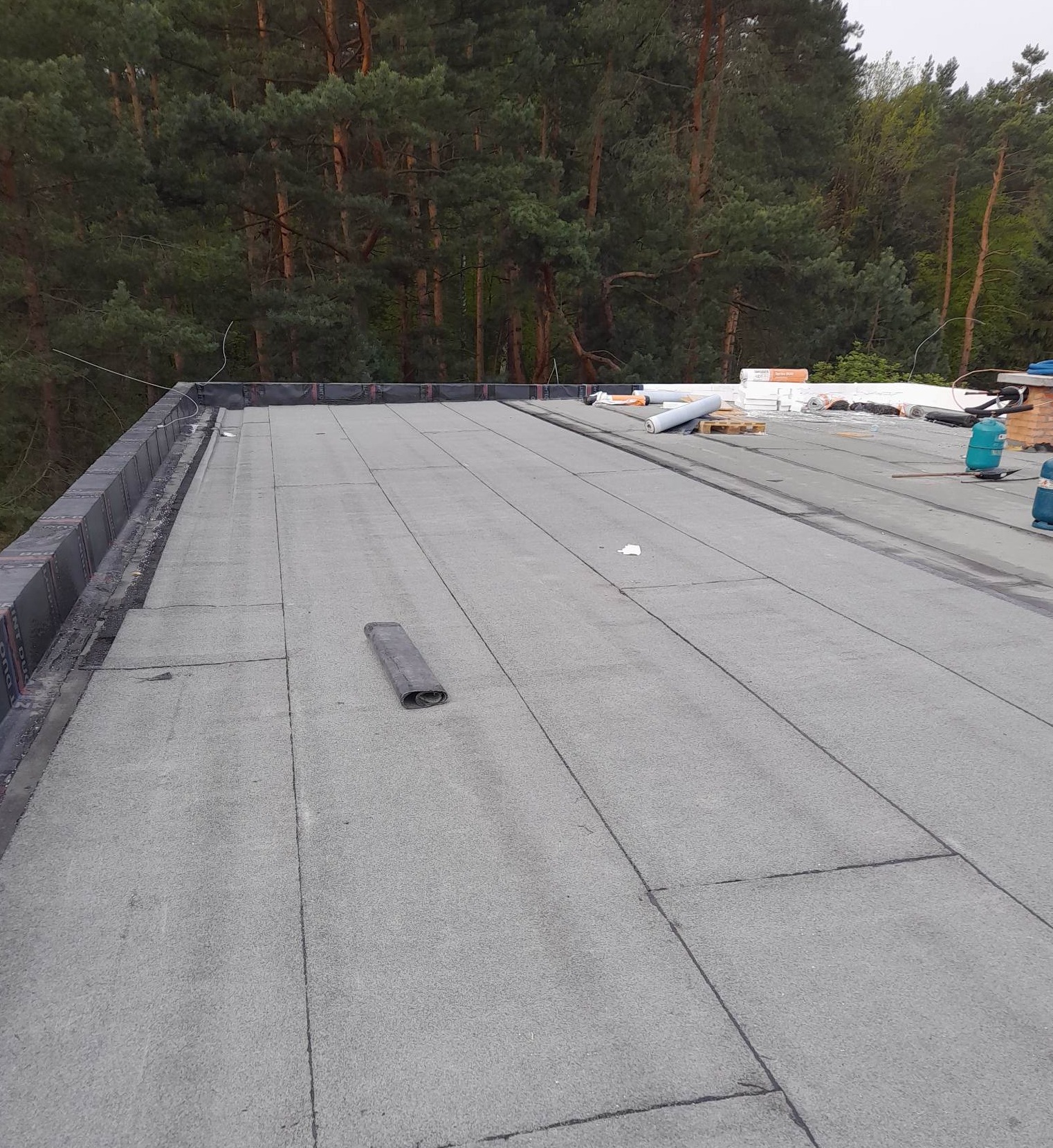 Płaski dach pokryty papą termozgrzewalną, widoczne zgrzewy, rolka papy, butle gazowe i narzędzia dekarskie w tle, drzewa w tle.