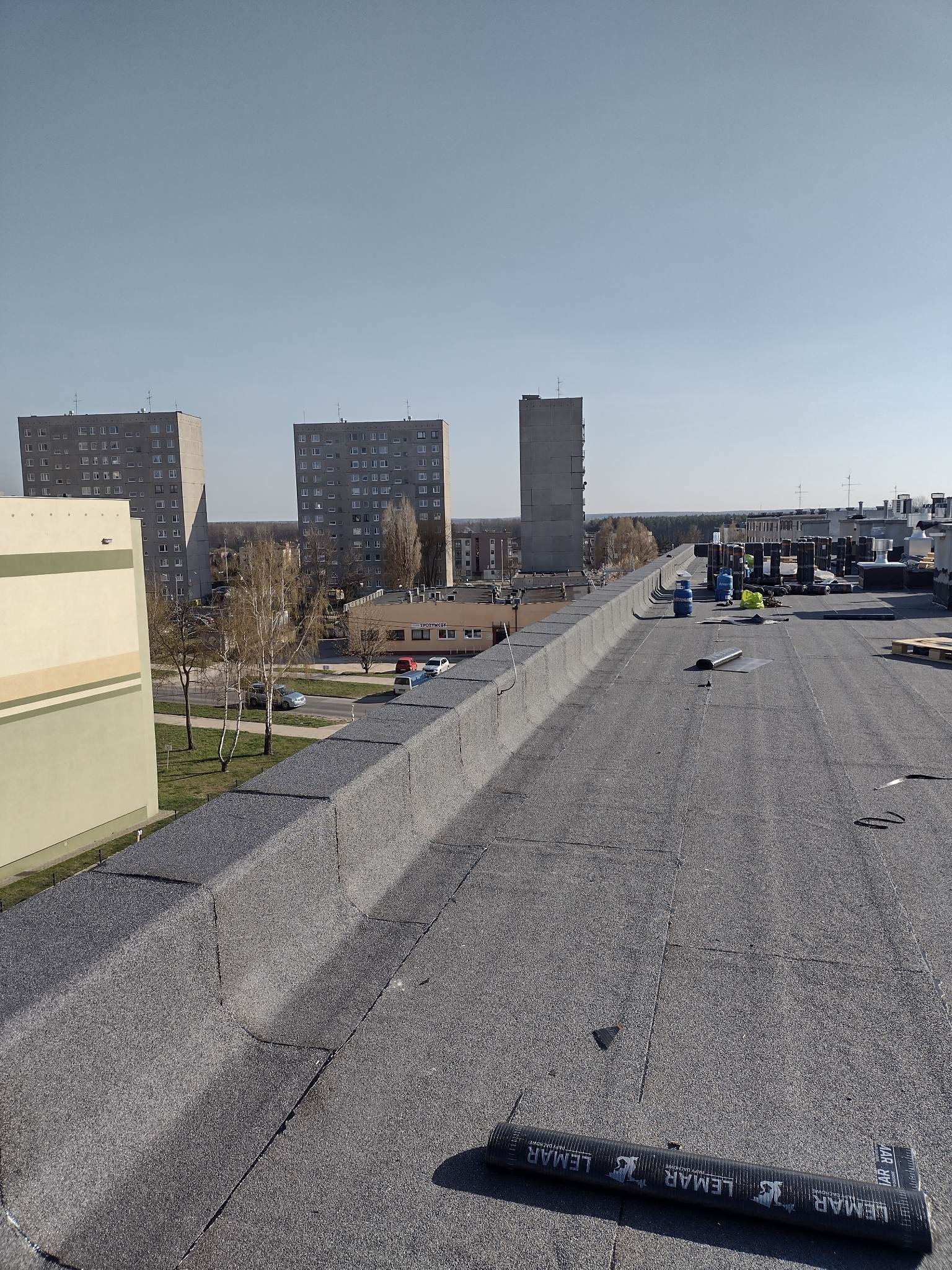 Widok z góry na płaski dach pokryty papą w Warszawie, z widocznymi blokami mieszkalnymi w tle i częściowo rozwiniętą rolką papy marki Lemar na pierwszym planie.