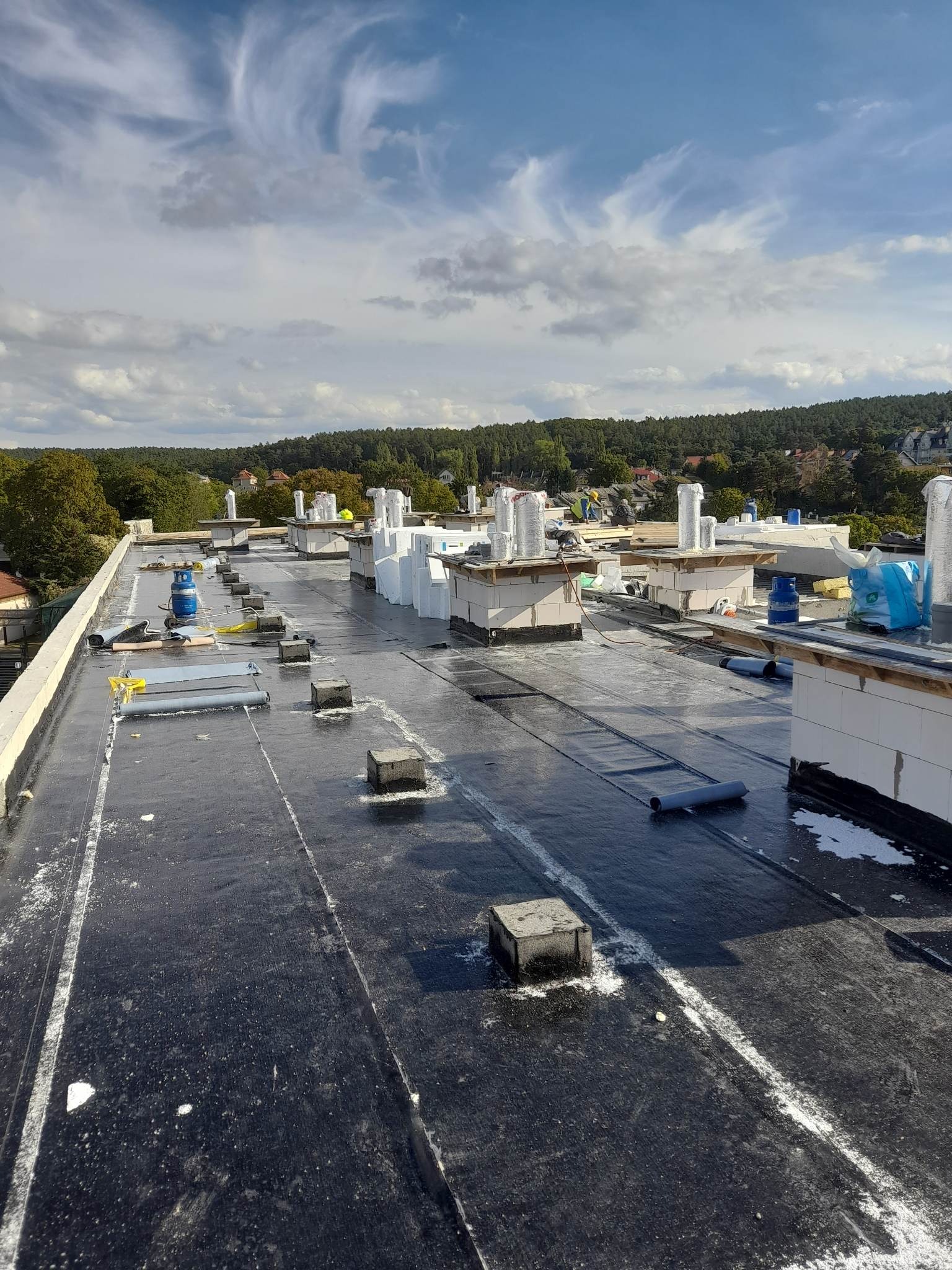 Czarny, płaski dach pokryty papą termozgrzewalną w trakcie prac, widoczne kominy wentylacyjne zabezpieczone folią, butla gazowa, rolki papy, bloczki betonowe, w tle zieleń drzew i zabudowania.