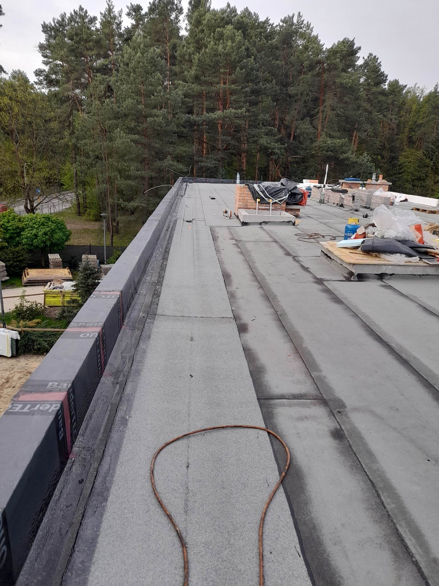 Płaski dach pokryty papą termozgrzewalną w trakcie prac, widoczne materiały izolacyjne przy krawędzi, przewody, komin w tle i las.