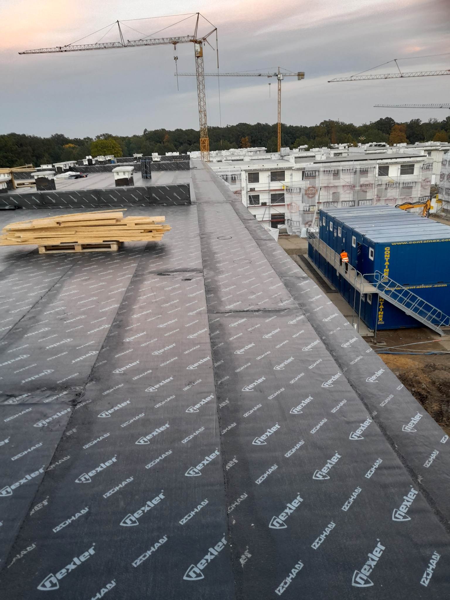 Widok z góry na płaski dach pokryty czarną membraną z widocznym logo producenta, na dachu leżą palety z drewnem, w tle budynki w trakcie budowy i żurawie budowlane.
