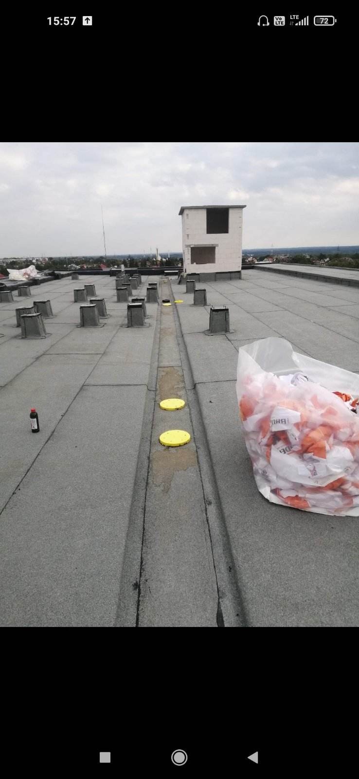 Płaski dach pokryty papą z elementami wentylacyjnymi, widoczny fragment rynny z żółtymi zaślepkami i workiem na śmieci, w tle zabudowa miejska.