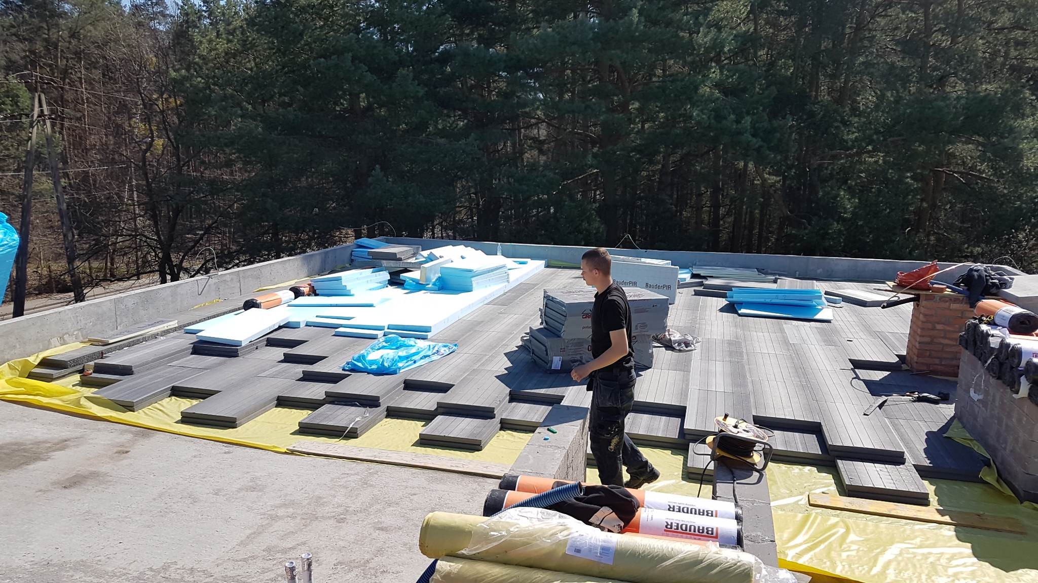 Prace na dachu płaskim: układanie izolacji termicznej z płyt styropianowych na folii, widoczny dekarz i materiały budowlane na tle lasu.