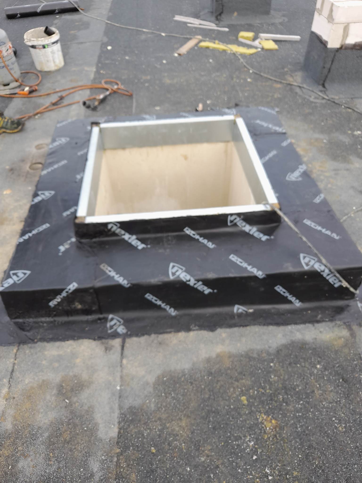 Montaż obróbki blacharskiej wokół otworu w dachu płaskim, zabezpieczonego membraną z nadrukiem, widoczne narzędzia i materiały budowlane w tle.