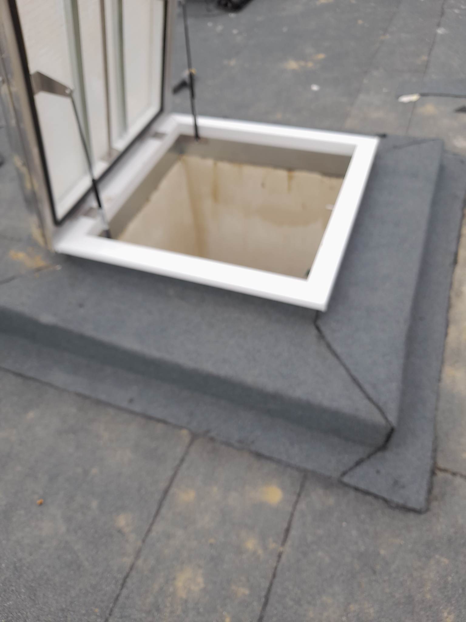 Otwarty właz dachowy z białą ramą, wbudowany w pokrycie dachu z papy bitumicznej, widoczne mocowania klapy włazu.