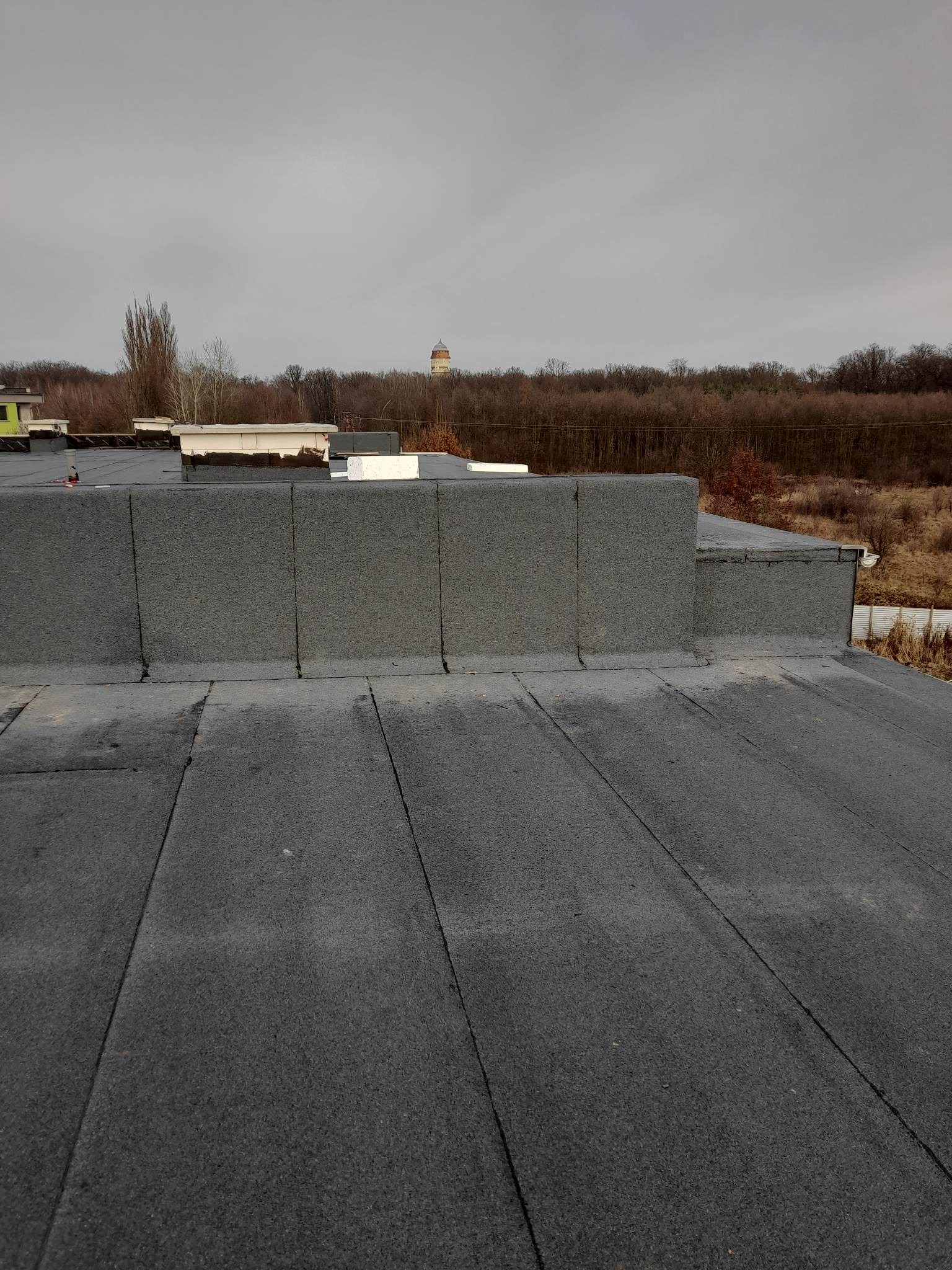 Widok z góry na płaski dach pokryty papą termozgrzewalną, z widocznymi kominami i elementami wentylacyjnymi, w tle drzewa i wieża ciśnień.