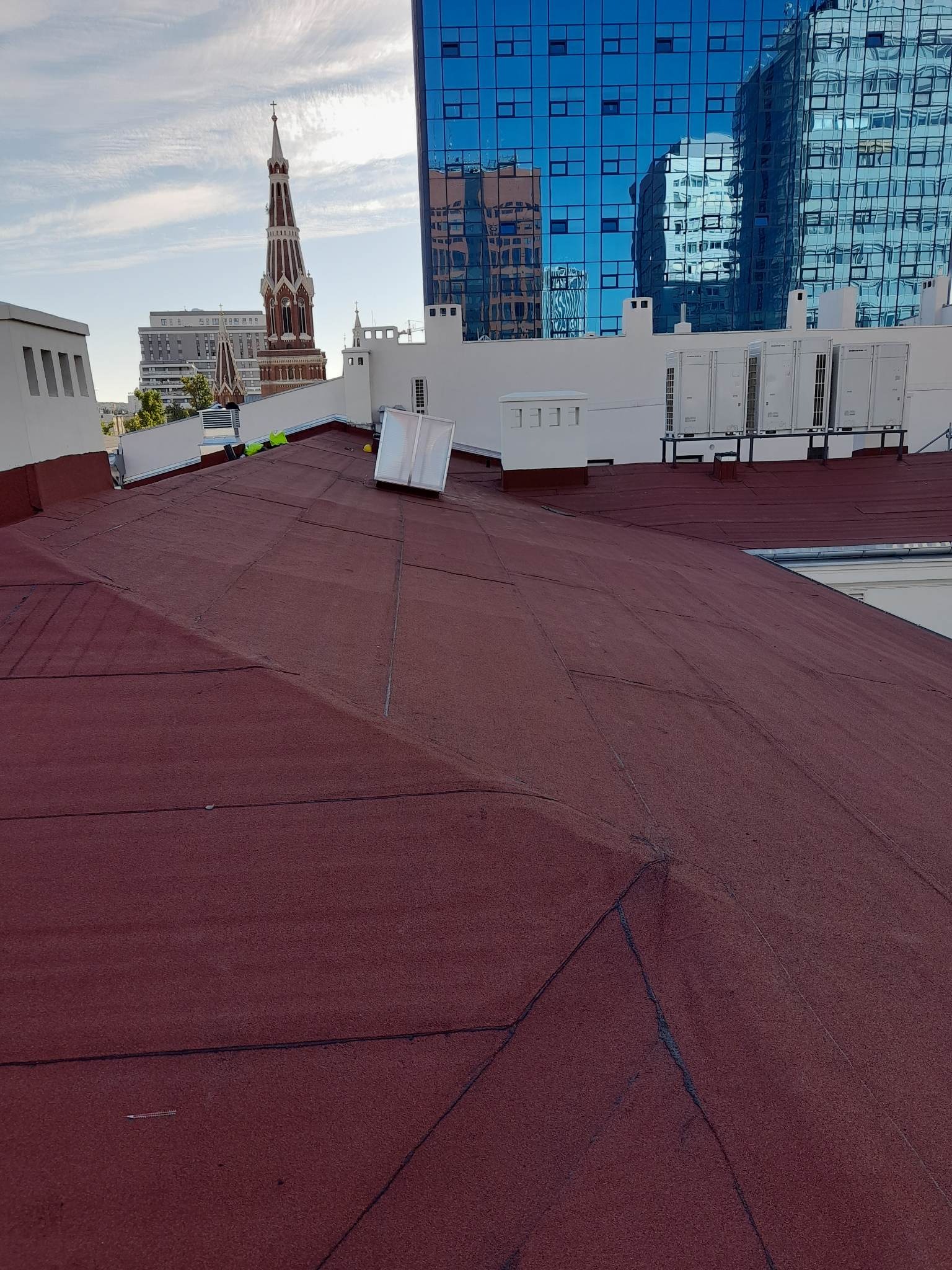 Czerwony dach pokryty papą na tle miejskiego krajobrazu z wieżą kościoła i nowoczesnym budynkiem biurowym, widoczne białe elementy wykończeniowe i klimatyzatory na dachu.