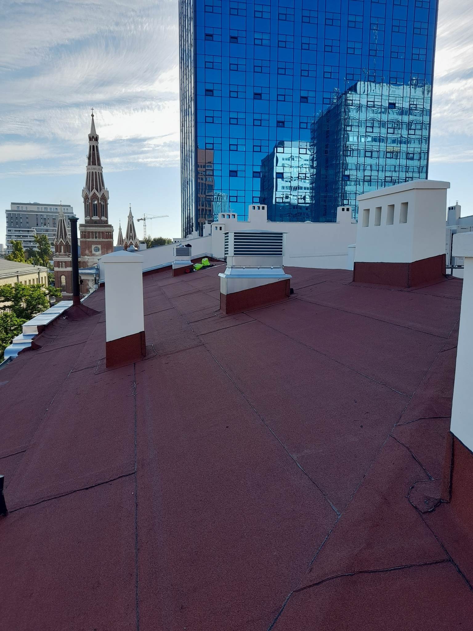 Ciemnoczerwony dach pokryty papą termozgrzewalną z widocznymi kominami i wentylacją na tle panoramy miasta z wieżą kościelną i nowoczesnym biurowcem.