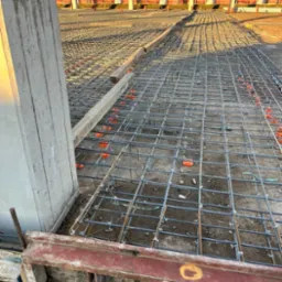 Przygotowanie do wylewki: siatka zbrojeniowa na podkładzie z dystansami, widoczne szalunki i fragment słupa konstrukcyjnego na tle budynku.