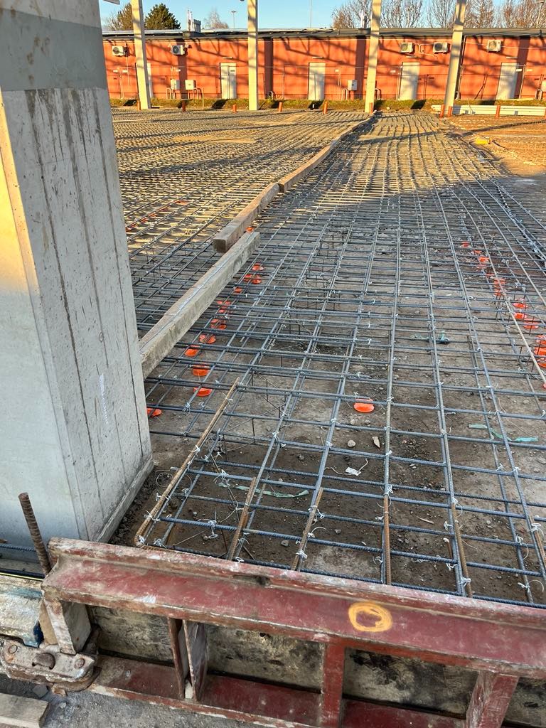 Przygotowanie do wylewki: siatka zbrojeniowa na podkładzie z dystansami, widoczne szalunki i fragment słupa konstrukcyjnego na tle budynku.