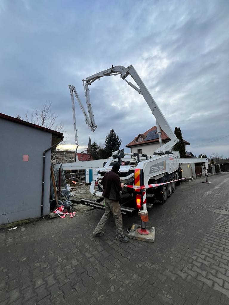 Pracownik obsługuje pompę do betonu Steeltech na chodniku z kostki, w tle budynek i pochmurne niebo.