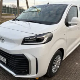 Biały, nowoczesny van Toyota z polską tablicą rejestracyjną PY 1754G, zaparkowany na ażurowej nawierzchni z widocznym logo Toyota na masce i reflektorami przednimi.
