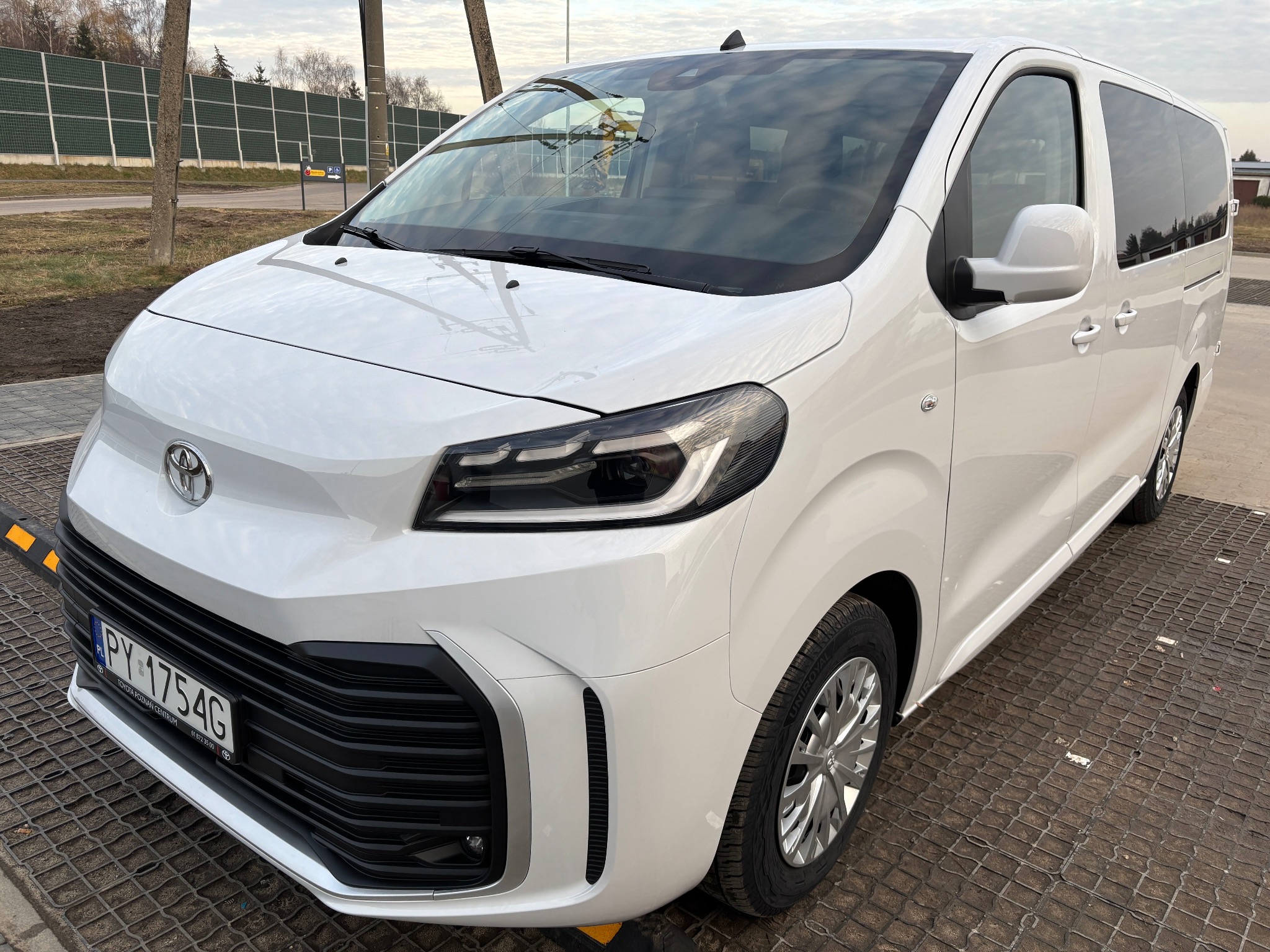 Biały, nowoczesny van Toyota z polską tablicą rejestracyjną PY 1754G, zaparkowany na ażurowej nawierzchni z widocznym logo Toyota na masce i reflektorami przednimi.