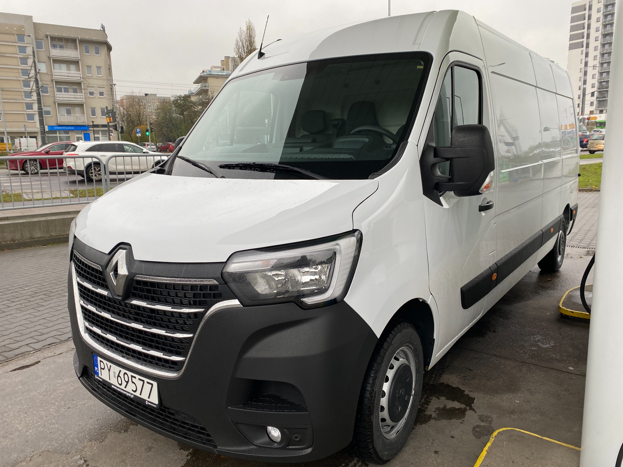 Biały samochód dostawczy Renault Master zaparkowany na stacji benzynowej, widok z przodu na mokrym bruku w pochmurny dzień.