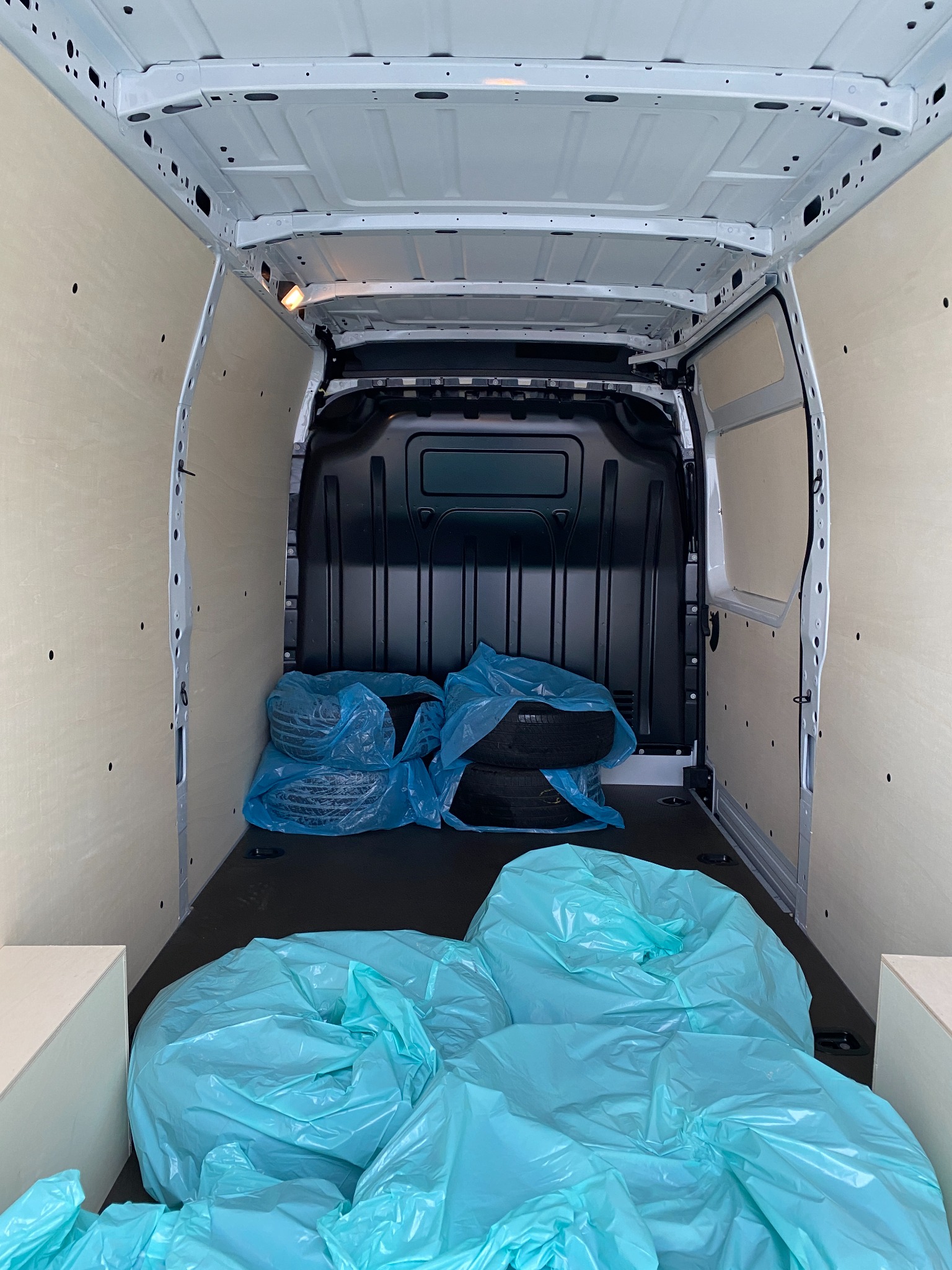 Wnętrze furgonetki z wyłożonymi sklejką ścianami i oponami w niebieskich workach foliowych.